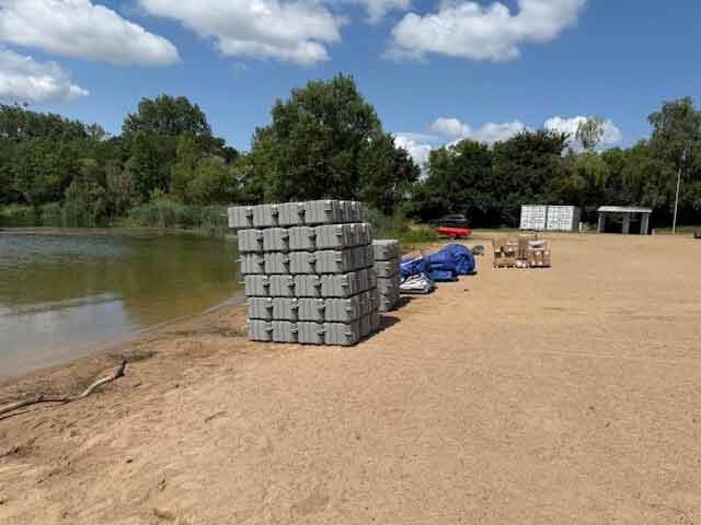 Palettes de flotteurs nautiques livrées par Cargomatic sur sable, préparation d’un ponton flottant sur plage au bord d’un lac.